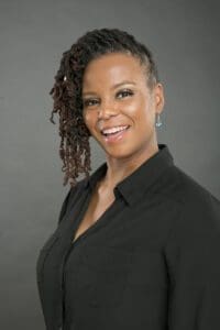 A woman with medium-dark skin and long, styled hair stands against a gray background, wearing a black collared shirt and smiling at the camera, embodying the professionalism often seen in nonprofit leaders. AlternativesBeauty.org