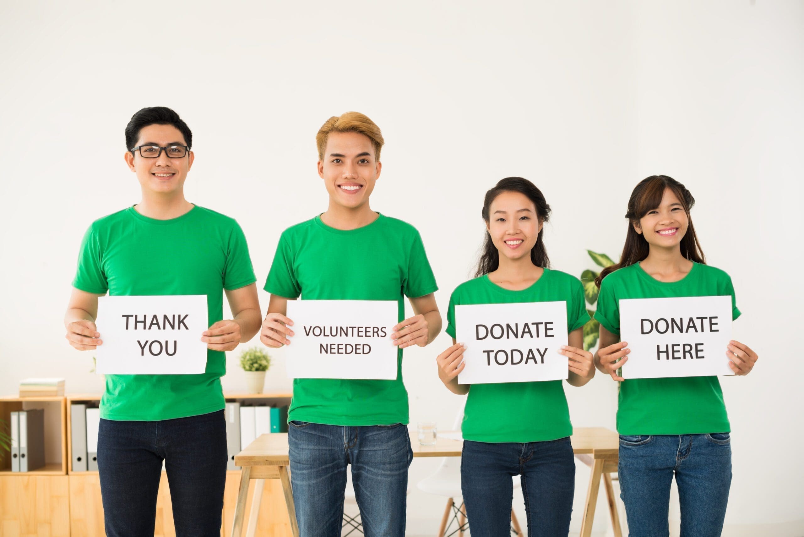 Four people in green shirts stand in a row, each holding signs that read: "THANK YOU," "VOLUNTEERS NEEDED," "DONATE TODAY," and "DONATE HERE" to support their nonprofit cause. AlternativesBeauty.org