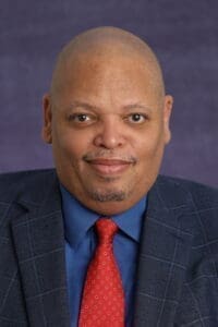 A man in a blue suit jacket, blue shirt, and red patterned tie poses for a formal portrait against a purple background, reflecting the professionalism often seen in nonprofit leadership. AlternativesBeauty.org