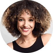 Woman with curly hair smiling, wearing a sleeveless black top, in front of a plain light background. AlternativesBeauty.org